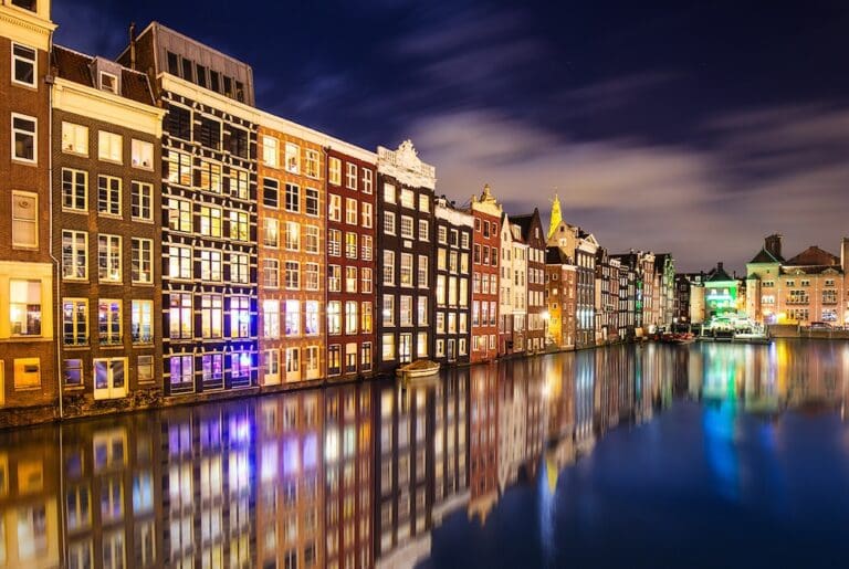 A nighttime view of a canal in Amsterdam with buildings in the background. The building lights reflect on the water